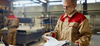 wp-680e583bc9e55c90-male-carpenter-wearing-face-mask-while-going-through-paperwork-workshop